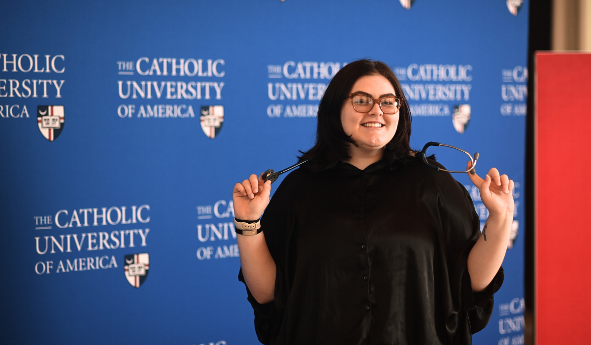 Olivia is standing in front of a blue Catholic University background. She has a stethoscope around her neck.