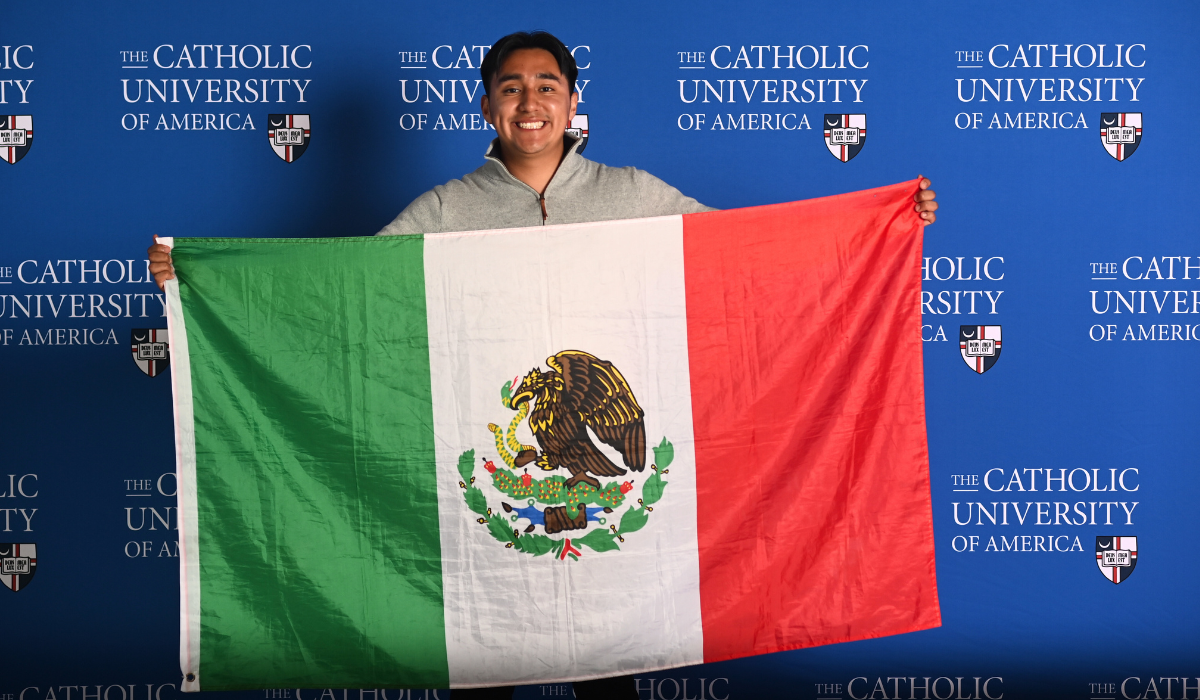 Luis Garcia Abundis stands in front of a blue Catholic University background while holding the Mexican flag.