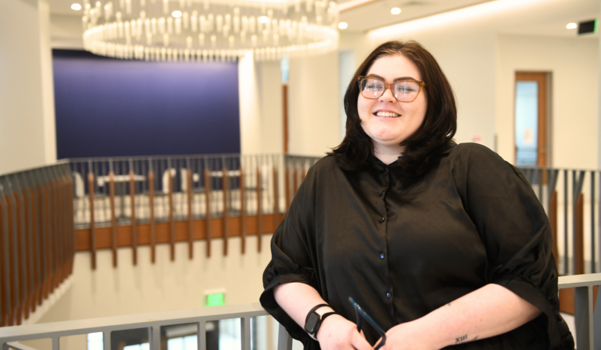 Olivia is standing in the foyer of The Conway School of Nursing. She is wearing a black button-down shirt. 