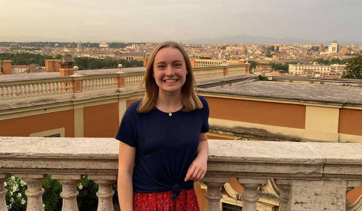 Elizabeth is standing in front of the skyline of Rome. She is wearing a black shirt and red pants. A silver necklace hangs around her neck.