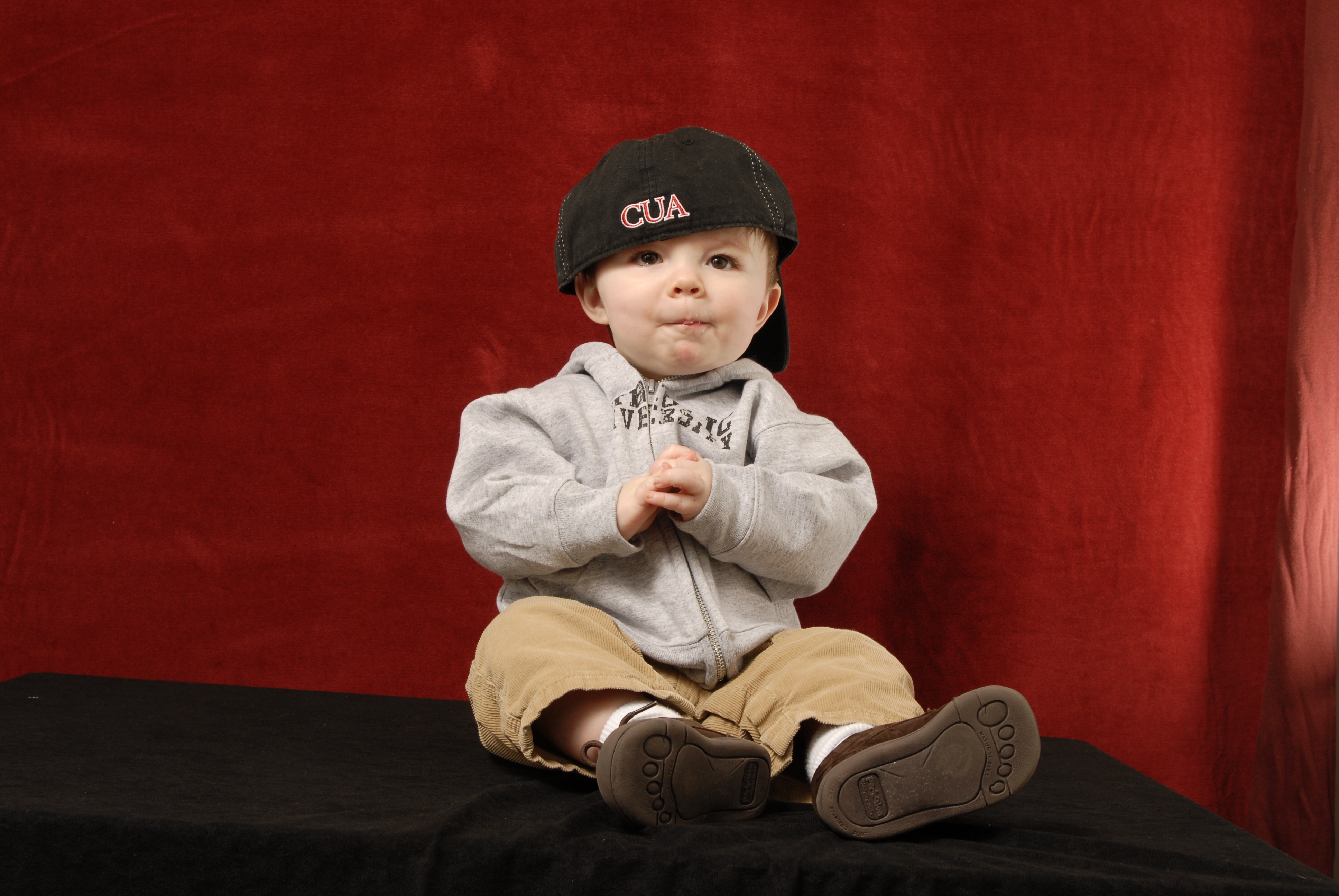 A toddler is dressed in a Catholic University sweatshirt and backwards baseball cap. He is sitting in front of a red backdrop.