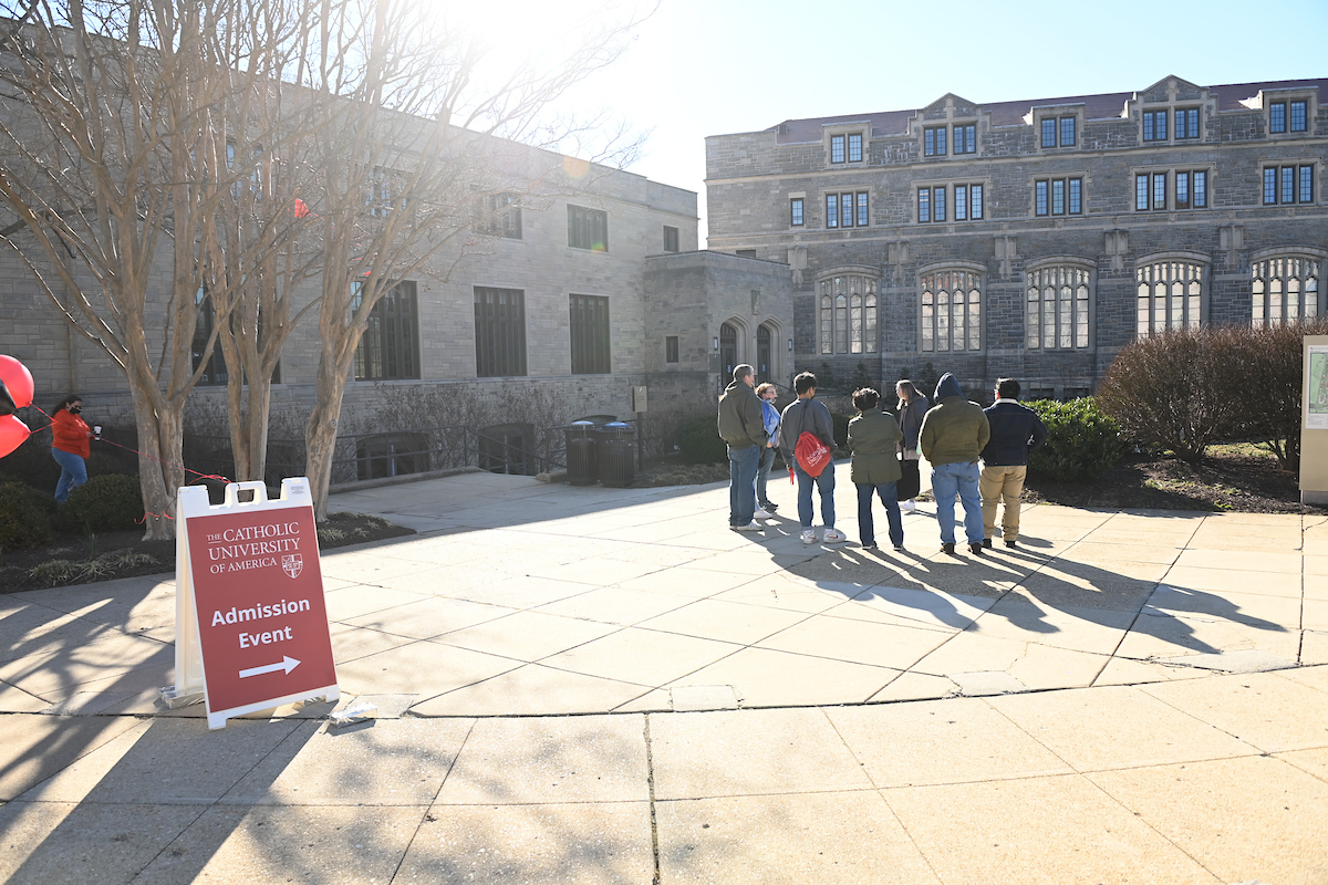 A group of recently admitted students and their parents are standing outside O'Connell Hall taking a tour of CUA campus. In the foreground, there is a sign welcoming Admitted Students with red and black balloons.