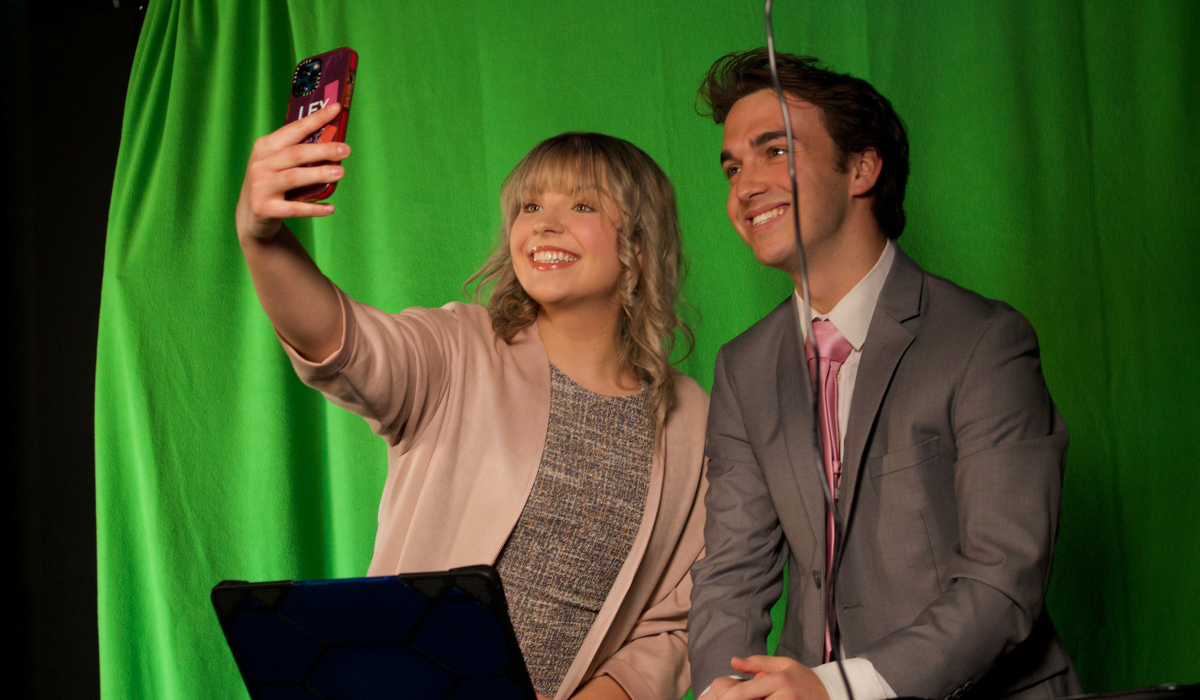 Alexis and a fellow student are taking a selfie in front of a green screen, preparing for a broadcast.