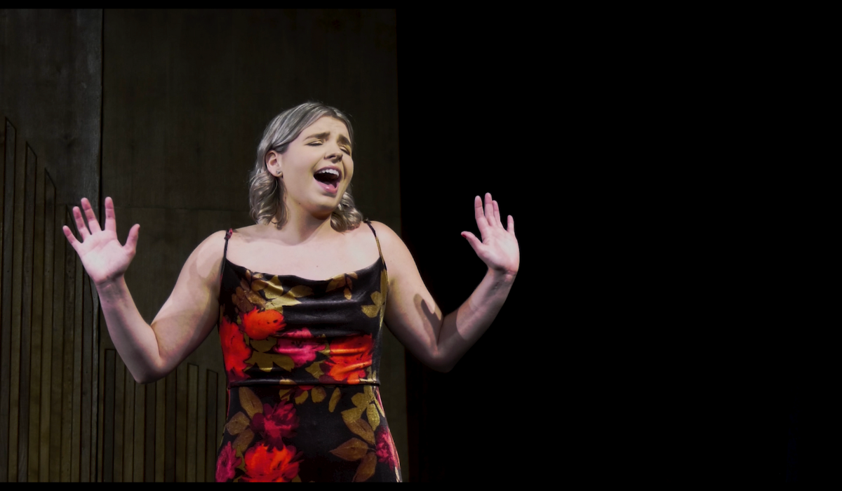 Alexis Greiss performs at a cabaret. She is wearing a dress with red and green flowers.