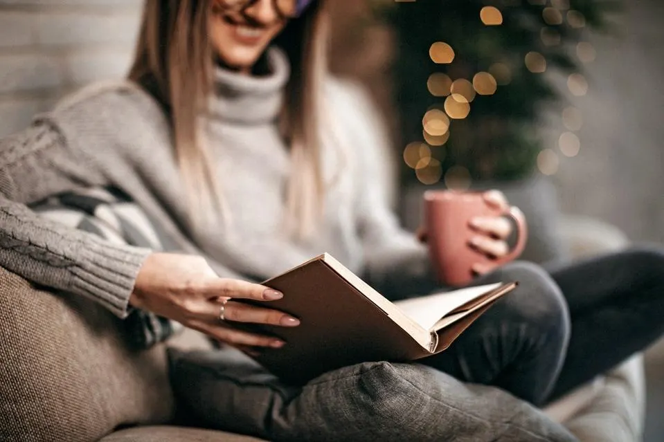 A woman is cozy in a winter scene, with the soft glow of a Christmas tree behind her. She is holding a book and a mug.