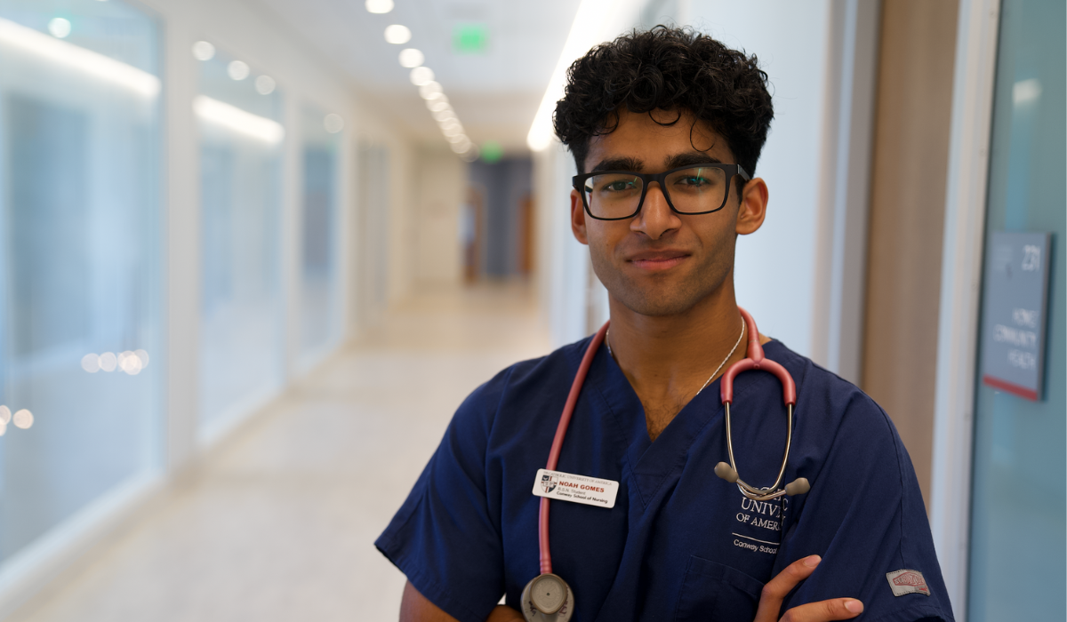 Noah Gomes is standing in the hallway of the Conway School of Nursing. He is wearing blue scrubs and a name tag. A stethoscope is draped around his neck.