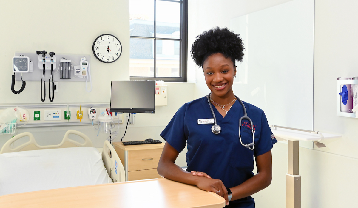 Anisa is in her dark blue scrubs in a hospital room. She has her stethoscope around her neck and is smiling. 