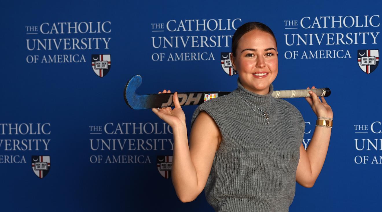 Mary Parker is standing in front of a blue Catholic University of America background. She has her field hockey stick draped over her shoulder.